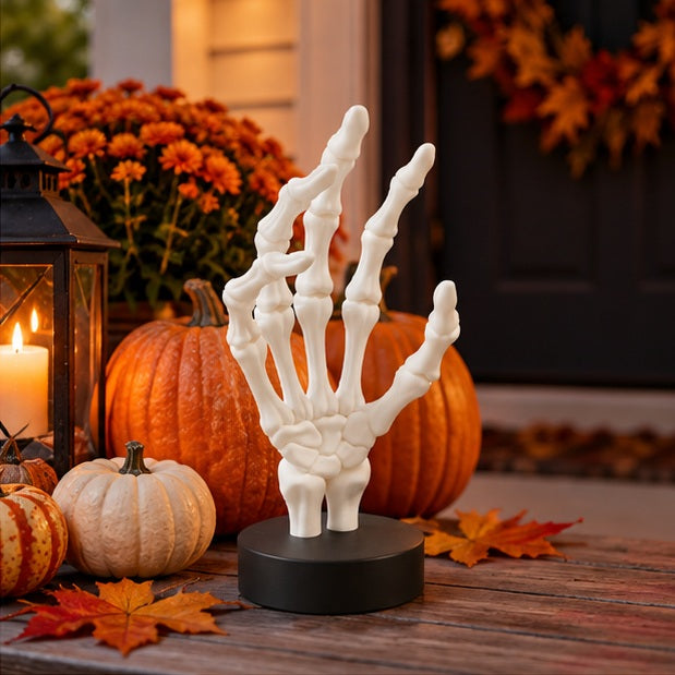 White skeletal hand on a black base in a spooky scene with a lantern, pumpkins and cobwebs